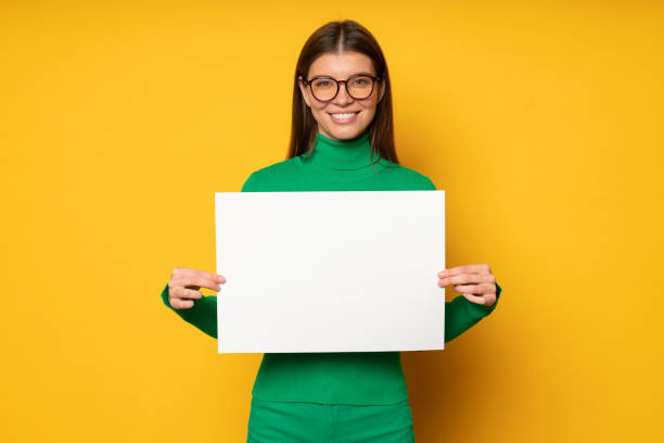 istock Woman in glasses posing on yellow background with white empty sheet with vacancy advertisement 1429310152
