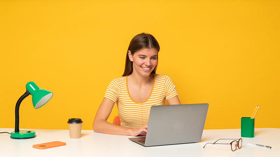 Friendly female freelance worker having video conference with her employer using laptop, sitting at her workplace on yellow studio background, checking e-mail