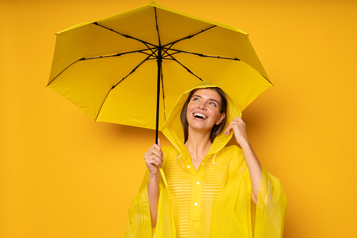 Happy dreamy woman in waterproof rain coat standing under umbrella looking up at cloudy sky with excitement, waiting for heavy rain, covering head with hood