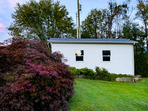 A side view of a tiny house. There is a Japanese Maple tree and lush green lawn in front of the house and trees in the back.  There is a small raised herb garden in front of the house that is lined with sandstone.  There is a utility pole behind the house.  A beautiful summer morning.  A Mother-In-Law home.