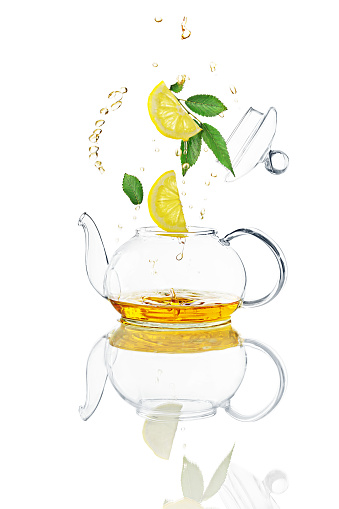 Flying green leaves, lemon slices, teapot lid, clear liquid splashes over transparent teapot. Concept of levitation, studio shot whith natural reflection, isolated on white.