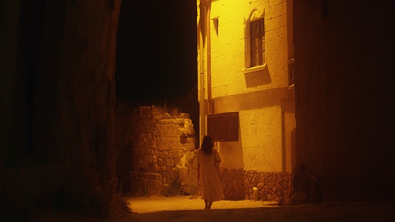 A young woman is walking in a small alley lit by a warm street light at night.