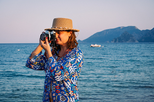 Young female photographer wearing a fedora taking pictures by the sea.