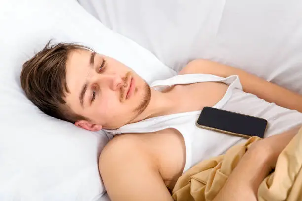 Sad Young Man with a Phone in the Bed at the Home