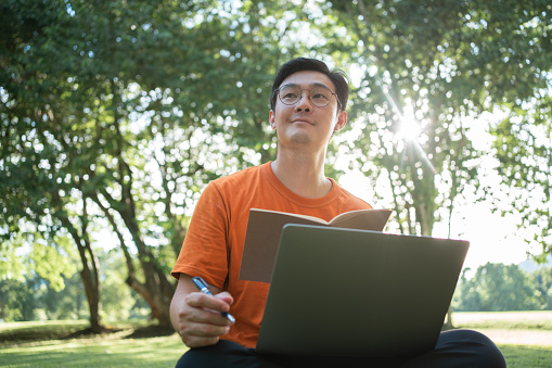 Southeast Asian Mature man student learning online class with Virtual Tutor and writing on a book. He studying and reskill on his work at the park. Wide shot