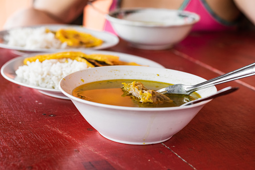 close-up of a typical Colombian dish of rib broth seasoned with natural spices and served on a red wooden table, in the background a peasant girl having breakfast before going to work.