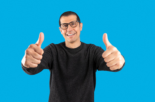 Studio shot of a man wearing casual sweater and glasses over blue background success sign doing positive gesture with hand, thumbs up smiling and happy. Cheerful expression and winner