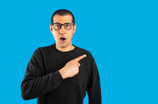 Studio shot of a hispanic man wearing casual sweater and glasses over blue background Surprised pointing with finger to the side, open mouth amazed expression