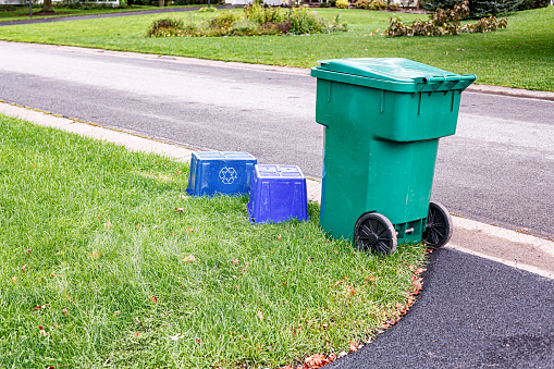 Empty, overturned blue recycling bins and an empty rolling garbage bin at the edge of a suburban residential district driveway and street intersection.