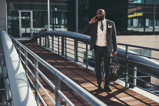 A mature fashionable black man entrepreneur is talking on the phone while passing on an overhead passage; an African bearded businessman is phoning while standing on the pedestrian bridge with his bag