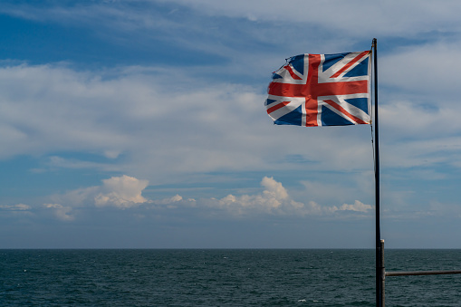 A tattered and torn Union Jack flag in a stiff wind with blue ocean and sky behind