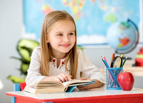 Little pretty blond girl sitting in the classroom at the white desk and reading book pointing by finger