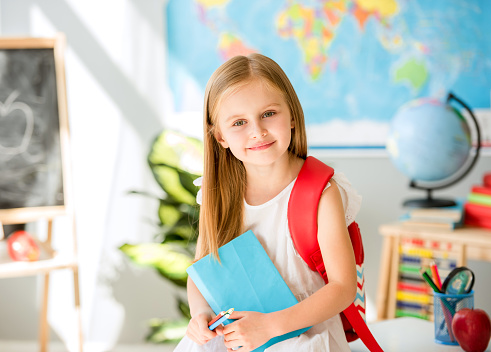 Little smiling blond girl standing near blackbord with red backpack and holding a notebook with pencils in the sunshine school classroom