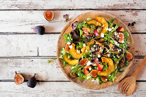 Autumn salad with roasted squash, apples, figs and feta cheese on a wooden plate. Overhead view table scene on a rustic white wood background.