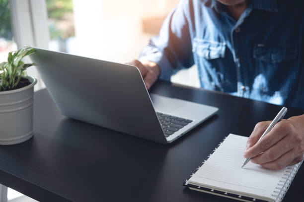 istock Close up of man working on laptop computer and hand with a pen writing on notebook on office table 1429409279