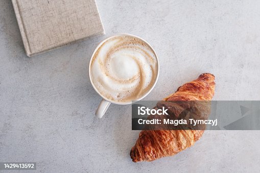 istock Cup of frothy cappuccino next to a croissant and a book 1429413592