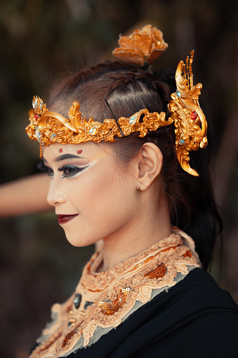 The face of an Asian woman with golden jewelry on her body while wearing a black costume with a happy face inside the jungle