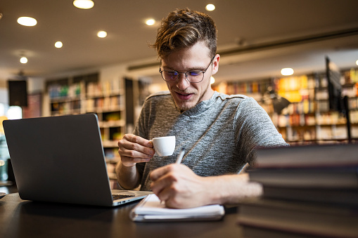 Young man sitting in modern bookstore ,library, reading and using his laptop