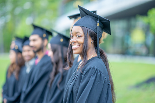 A group of University students stand in a line outside on graduation day as they pose for a portrait.  They are each dressed formally in a gown and cap as they smile and enjoy the success of their hard work.