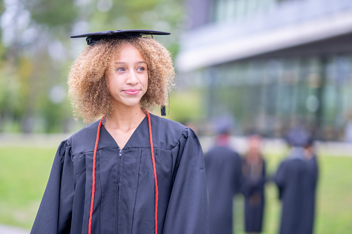 A female University graduate, of African decent, poses for a portrait outside on campus.  She is dressed formally in a gown and cap as she poses for a graduation photo.