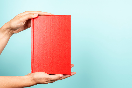 Woman hands holding book with blank red cover over light blue background. Education, back to school, self-learning, book swap, sharing, bookcrossing concept.