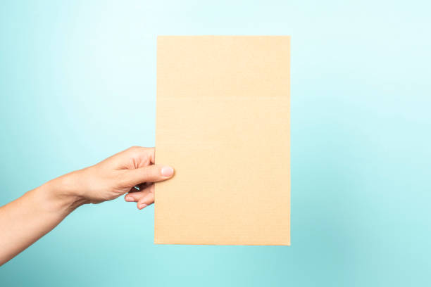 istock Woman hand holding blank rectangular sheet of brown cardboard on light blue background 1429434535
