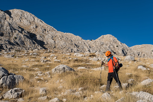 60 years old man. He is trekking. Mountainous extreme region. Orange t-shirt, backpack, walking pole, helmet.Steep mountain in blue clear sky background.