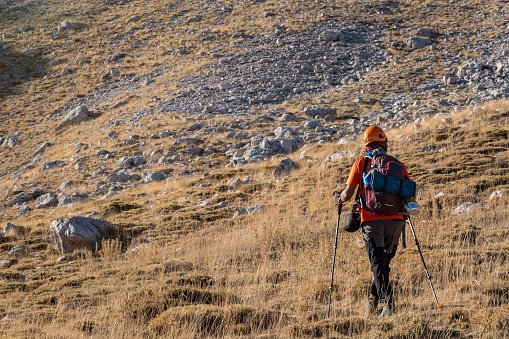 Aka terrain full size model. Hiking in extreme rocky terrain.60 years old man. He is trekking. Mountainous extreme region. Orange t-shirt, backpack, walking pole, helmet