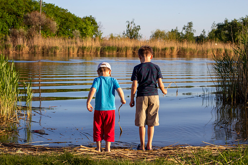 Young Hispanic and African American fishing buddies walk to the lake or pond to fish together. An Hispanic boy is carrying a fishing pole and tackle box. An African American boy is holding a fishing pole and net in the background.