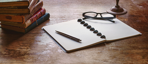 An open notebook with a pen and glasses on an old wooden table with a stack of vintage books on it. Wide panoramic image with copy space for template and background