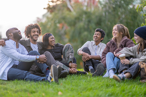 A small group of young adults sit in the grass together as they spend time together socially.  They are each dressed casually in fall layers and are laughing as they enjoy the fresh air and time together.