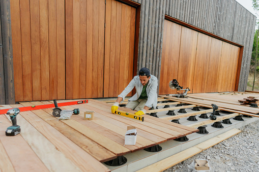 Photo of a carpenter using a level while attaching wooden decking boards to a deck of a house.