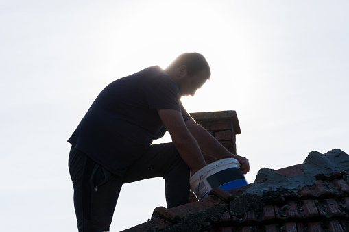 Worker secure and patch roof tiles with cement on an old building roof in the village on a sunny day. Silhouette of a man on a roof while working