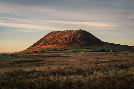 Slemish mountain on a summer evening at sunset, with beautiful dusk side lighting and overcast sky. The grass fields are bordered in distance with hand built stone walls. The mountain is a popular tourist attraction, and for walking. St. Patrick tended sheep there as a young man. The mountain is actually the plug of an extinct volcano