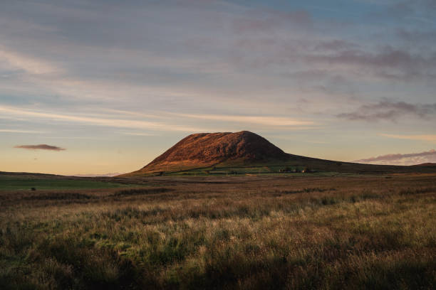 Wide view of Slemish mountain at dusk, at County Antrim, Northern Ireland Slemish mountain on a summer evening at sunset, with beautiful dusk side lighting and overcast sky, shot in wide angle. The grass fields are bordered in distance with hand built stone walls. The mountain is a popular tourist attraction, and for walking. St. Patrick tended sheep there as a young man. The mountain is actually the plug of an extinct volcano extinct volcano stock pictures, royalty-free photos & images