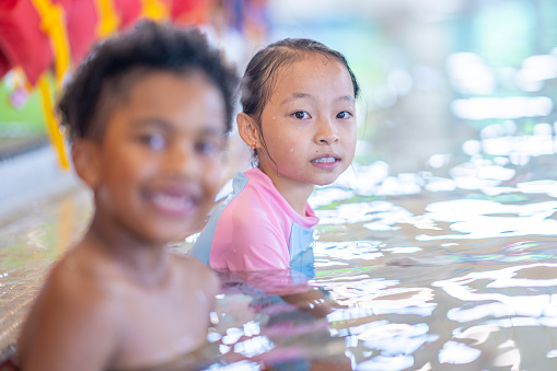 A young girl of Asian decent smiles at the camera as she sits in the pool beside a male friend during a swimming lesson.  Lifejackets can be seen hanging in the background at the pools edge.