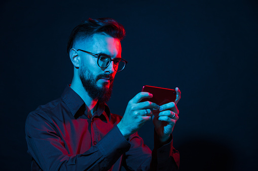 Studio portrait of a white 30 year old bearded man in glasses with a mobile phone on a black background