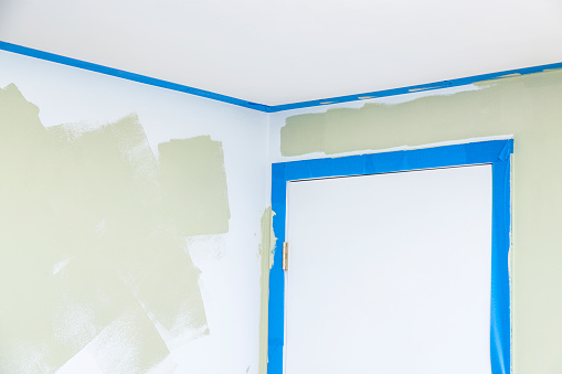 A home bedroom wall has been partially painted using a paint roller. The closed white bedroom door on the right and the ceiling both have blue painters' masking tape to separate and protect those areas from the new paint.