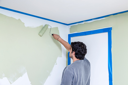 Right handed young man re-painting a home bedroom wall using a paint roller. The closed white bedroom door on the right and the ceiling both have blue painters' masking tape to separate and protect those areas from the new paint.