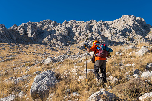 60 years old man. He is trekking. Mountainous extreme region. Orange t-shirt, backpack, walking pole, helmet.Steep mountain in blue clear sky background.