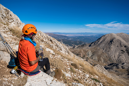 60 years old man. He is trekking. Mountainous extreme region. Orange t-shirt, helmet.Steep mountain in blue clear sky background.The model sits on the stone and opens the lid of the flask to drink water.