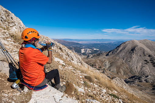 Man drinking water against the view. Space for text.60 years old man. He is trekking. Mountainous extreme region. Orange t-shirt, helmet.Steep mountain in blue clear sky background.The model sits on the stone and opens the lid of the flask to drink water.