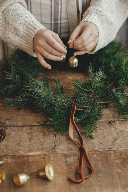 Making Christmas rustic wreath. Woman hands holding golden bells and decorating wreath on rustic wooden table with ribbon, cones, candles. Moody holiday image. Winter holiday workshop stock photo