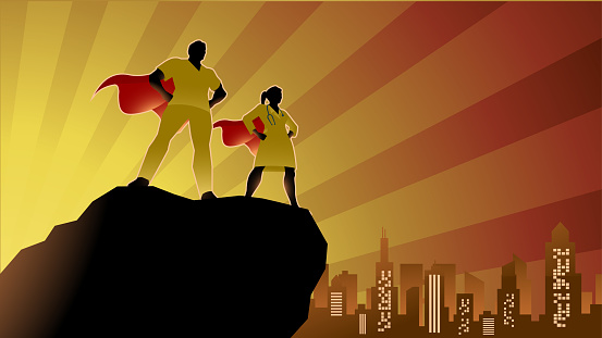 A silhouette style vector illustration of a couple of doctors or medical workers depicted as superheroes standing on a cliff with city skyline and sunburst in the background. Wide space available for your copy.