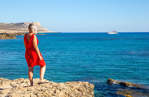 View from back of slender girl in red dress on edge of cliff overlooking Mediterranean Sea