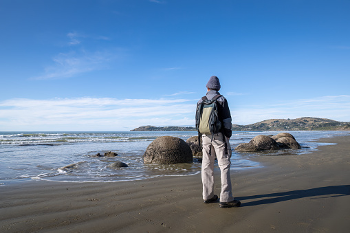 Tourist looking at waves splashing around the boulders at Moeraki Boulders beach, Otago.