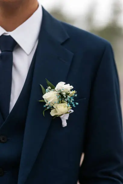 Close-up of the groom's wedding boutonniere of cream eustoma or rose flower and green leaves pinned to a blue jacket against a background of a white shirt and tie. Wedding accessory.