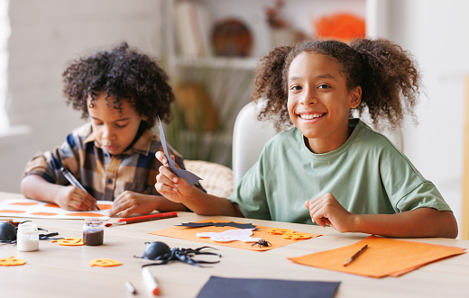 Happy african american children girl and boy making Halloween home decorations together, kids painting pumpkins and making paper cuttings