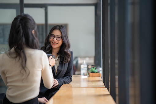 Young beautiful Asian businesswoman having fun talking with her colleagues during a break from work. Asian businesswoman wearing glasses sharing ideas, discussing the project with a colleague.