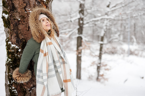 Portrait of young woman leaning on tree, looking at view\n and smiling in snowy forest on cold winter day